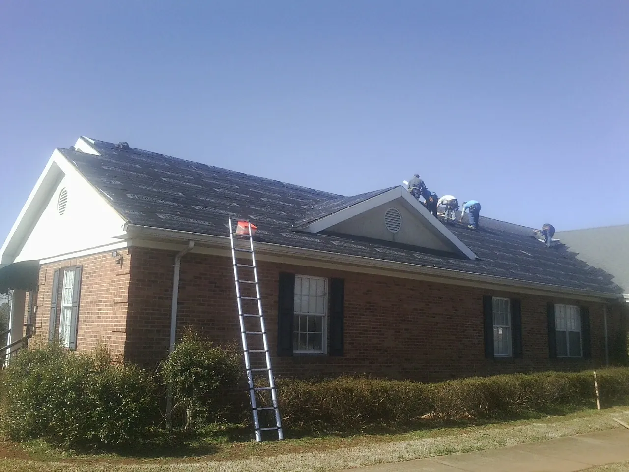 Crew tearing shingles off a brick ranch roof with a ladder set — Metal Roof Repair work by our crew in Manorville