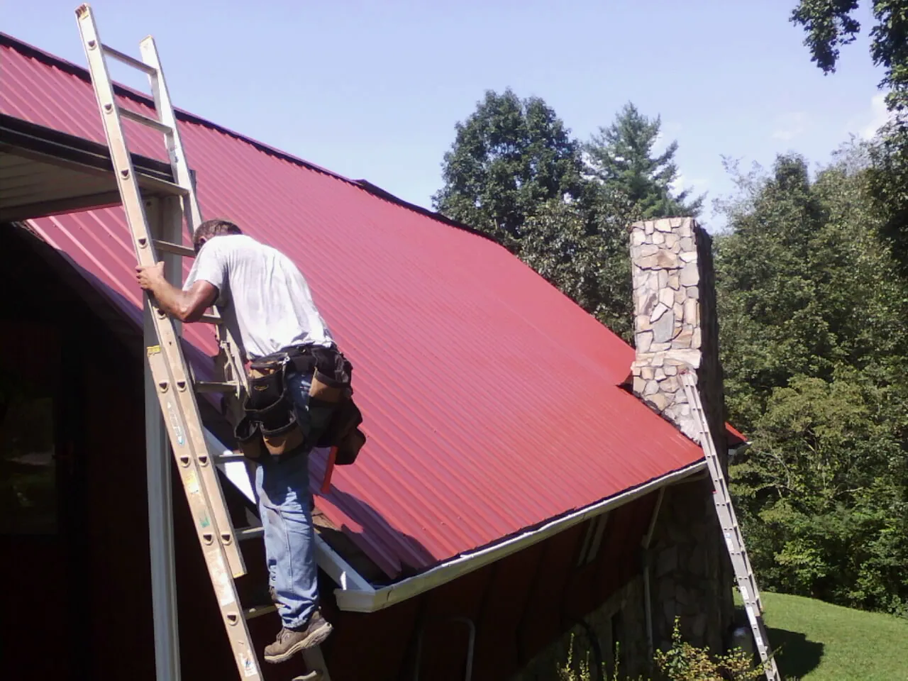 Roofer on a ladder beside a red metal roof and stone chimney — Soffit & Fascia Repair by Roofing Brothers in Manorville